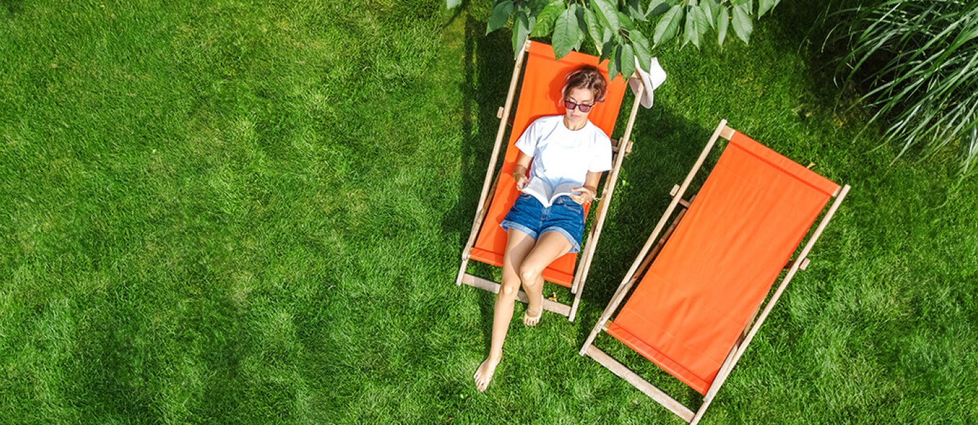 Femme qui lit étendue au soleil dans un jardin avec une belle pelouse dans une cour aménagée.